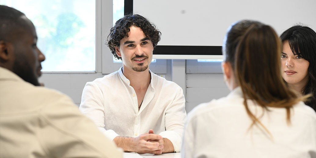 étudiant cheveux bruns bouclés en chemise blanche qui sourit à côté d'une étudiant avec les cheveux lisse noir. Devant eux deux étudiants dos à la caméra