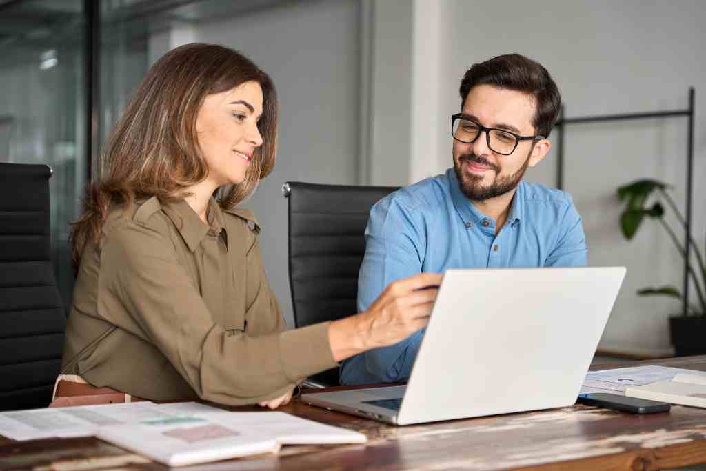 Une femme brune en chemise verte discute avec un homme brun avec des lunettes en chemise bleu dans un bureau devant un ordinateur portable